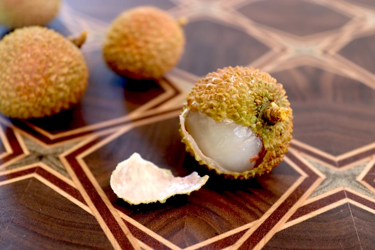 Lychee fruit with a bit of the skin peeled off to reveal the flesh, on a dark wood cutting board.