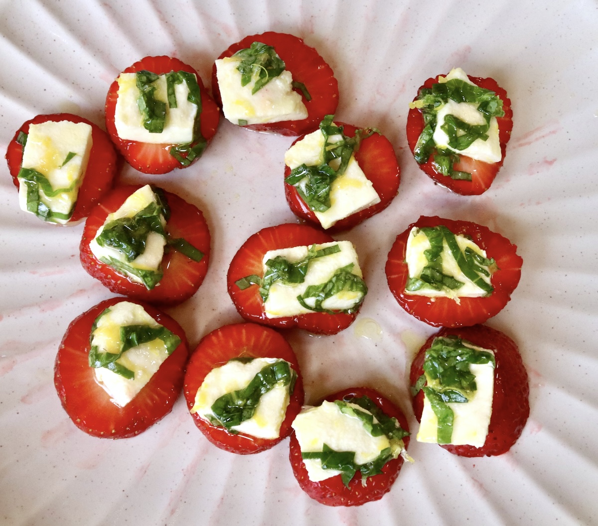 12 slices of fresh strawberries with pieces of fresh mozzarella and chopped basil, on a white plate.
