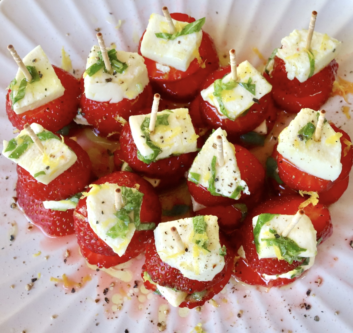 12 tiny stacks of strawberry caprese with toothpicks, on a white crimp-edged plate.