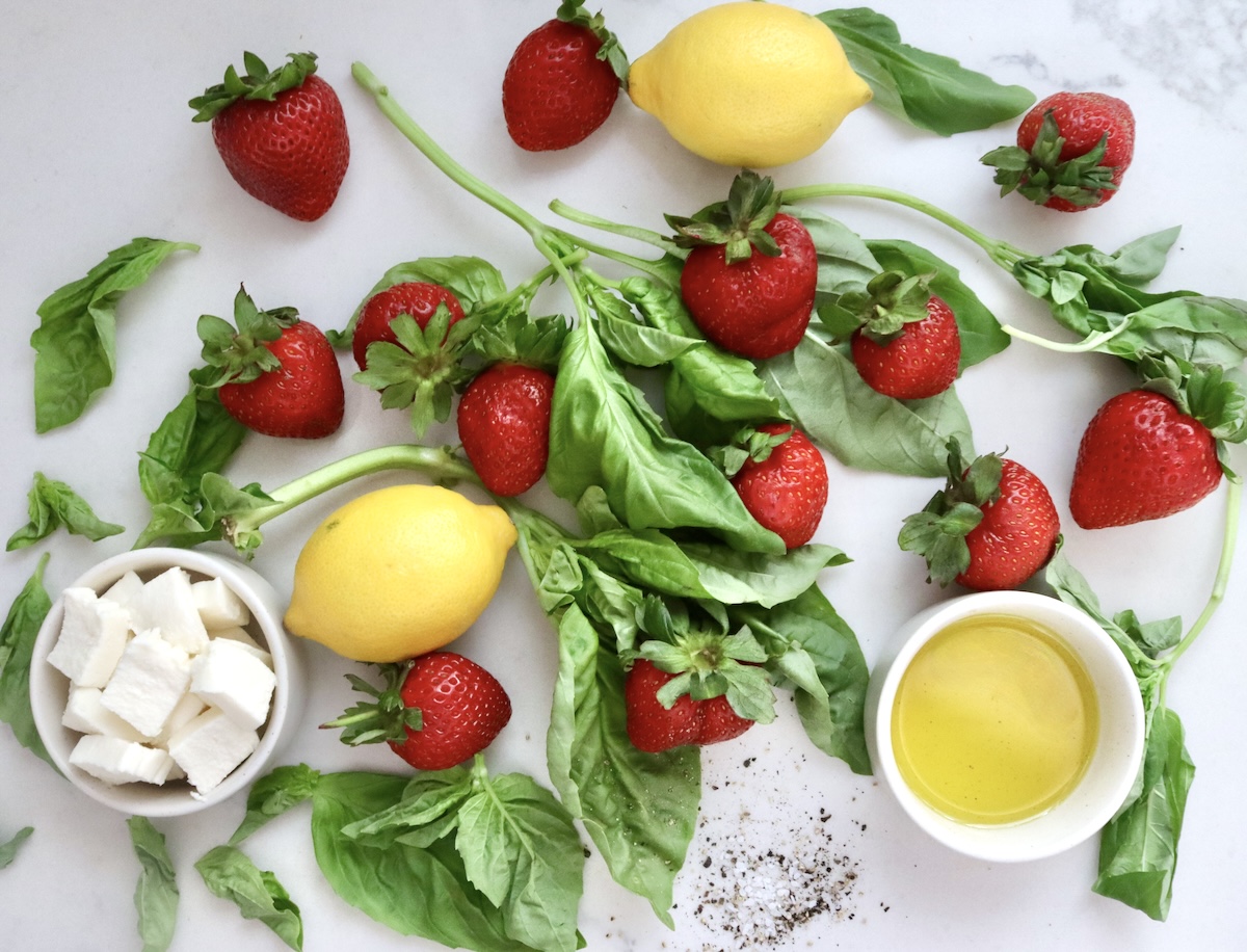 Strawberry Caprese Appetizer ingredients on white marble, including olive oil, lemons, fresh mozzarella, fresh basil, salt and pepper.