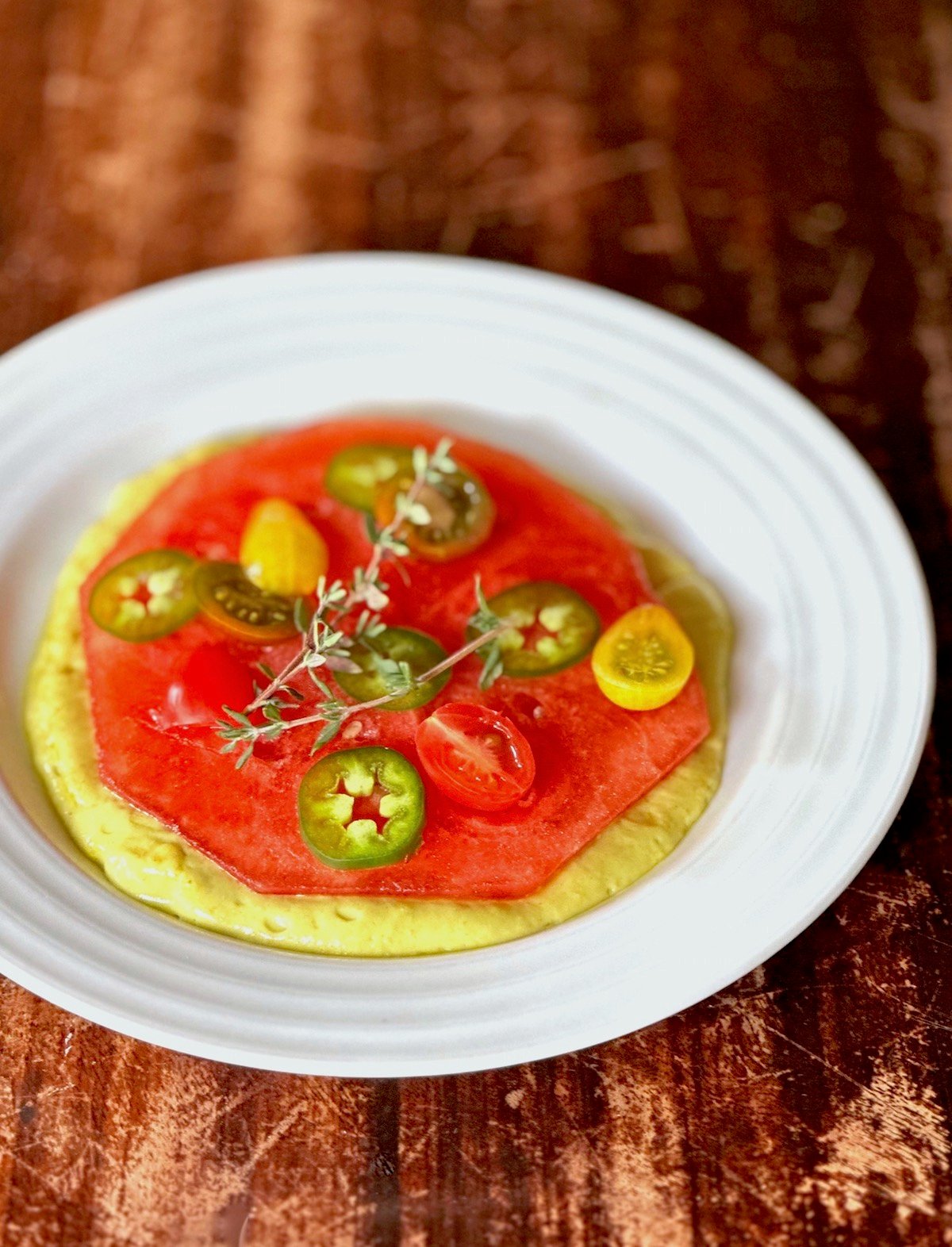One round piece of compressed watermelon on a white plate with jalapeno and tomato slices and sprig of thyme.
