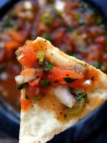 Tomato Poblano Salsa on a tortilla chip held above a dark grey bowl filled with more salsa.