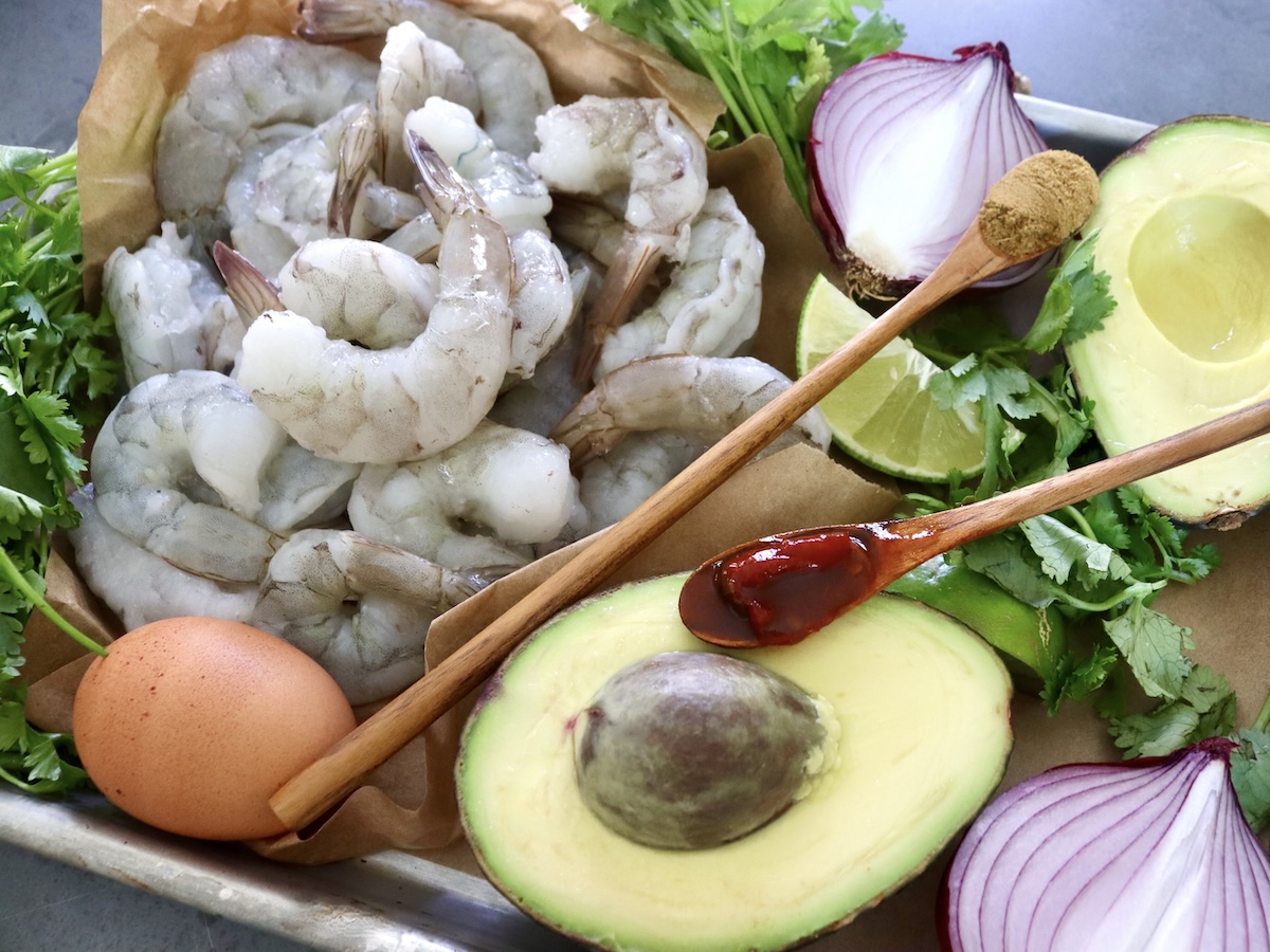 Ingredients for shrimp burgers with guacamole including raw shrimp, 1 egg, red onion, cilantro, lime, cumin in a wood spoon and chipotles in a wood spoon.