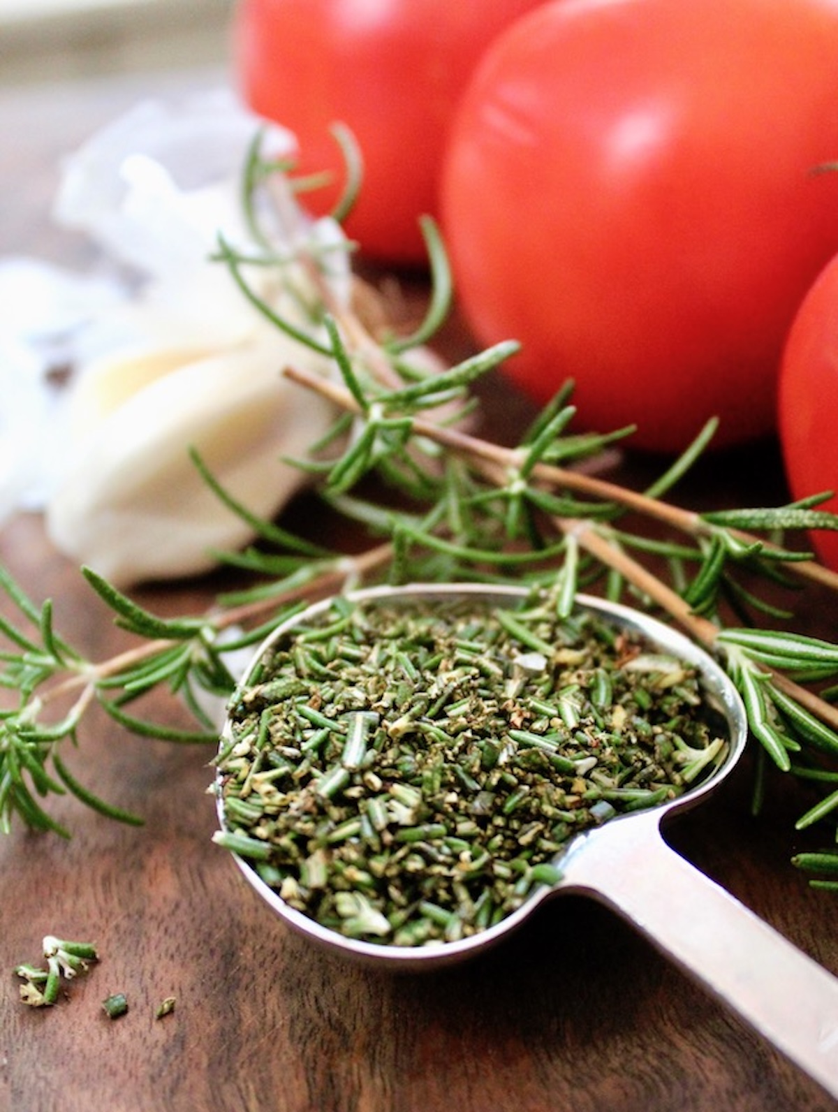 Heart-shaped tablespoon with chopped rosemary, garlic cloves and fresh tomatoes, on a cutting board.