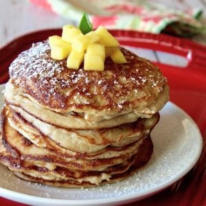Tall stack of coconut flour pancakes on a white plant that's on a red tray, with pineapple on top.