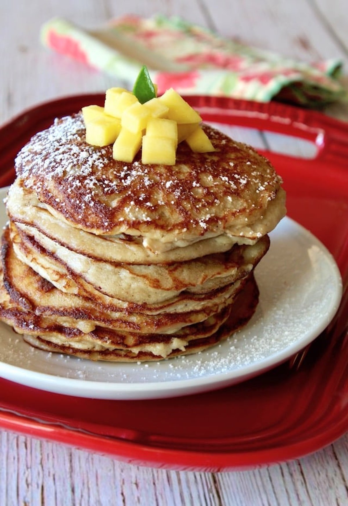 Tall stack of coconut flour pancakes on a white plant that's on a red tray, with pineapple on top.
