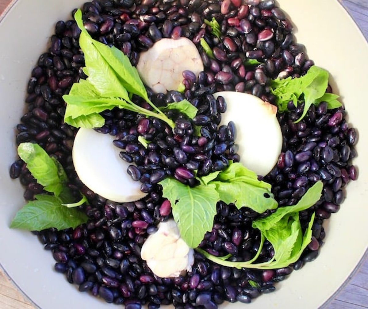 Large pot with soaked black beans with chunks of onion, head of garlic and green epazote leaves.