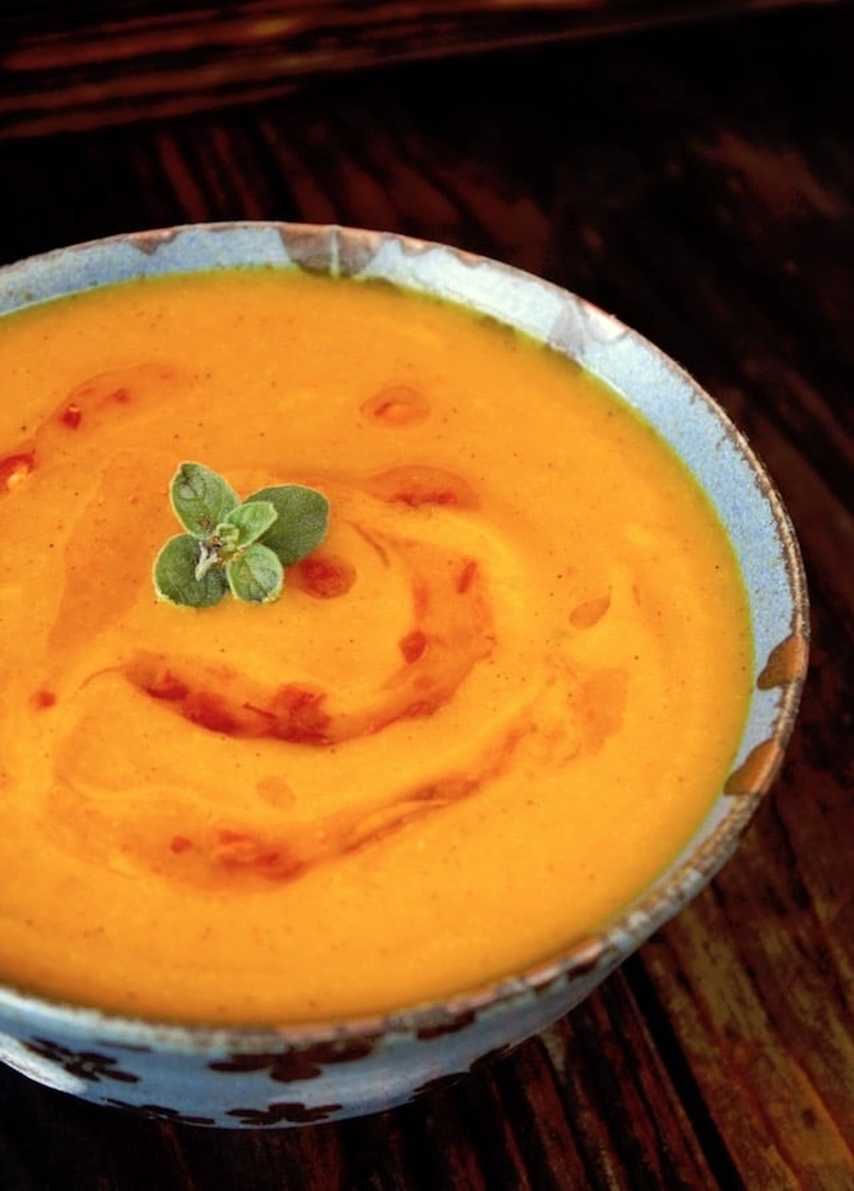 Top view of bright orange Red Kuri Squash pureed soup, in a blue bowl with a small oregano leaf on top.