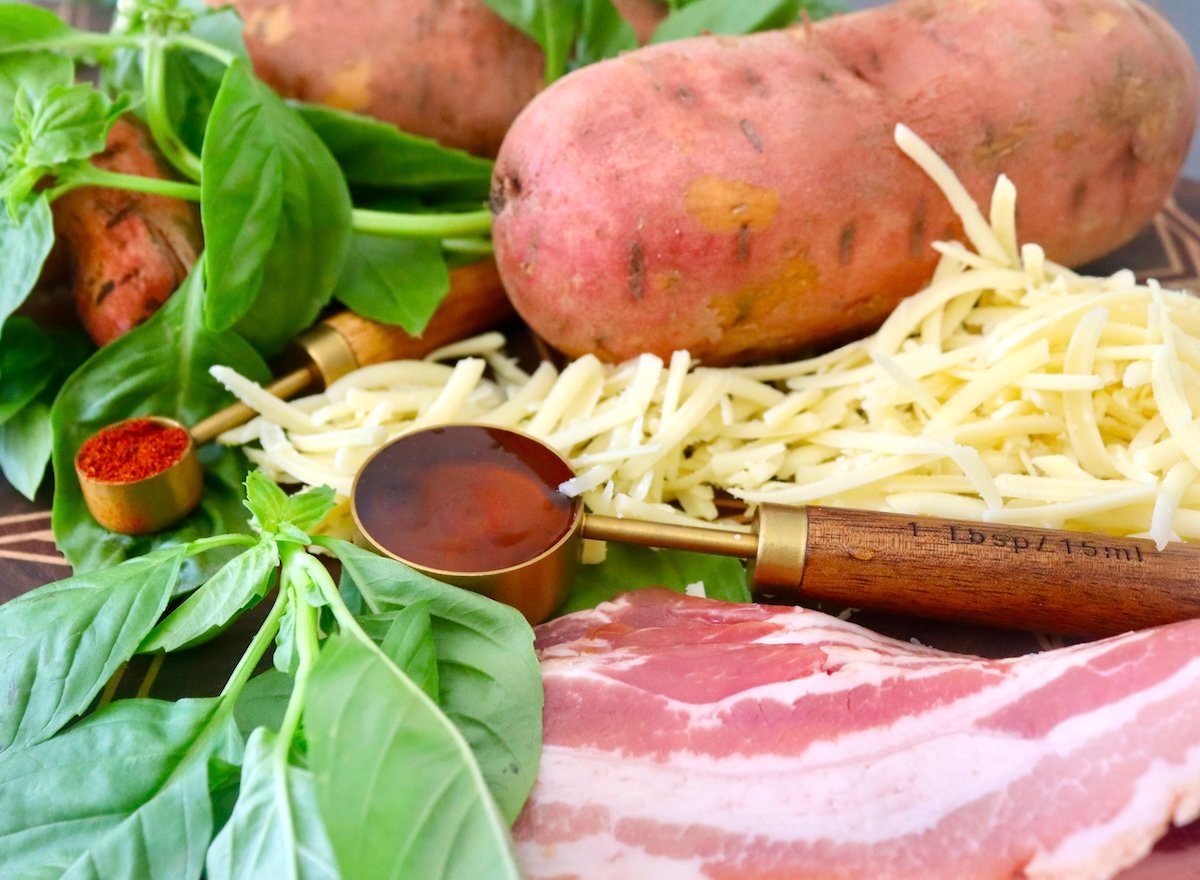 Grated white cheese, 3 whole raw sweet potatoes, raw bacon slices, honey and chili powder in a measuring spoon.