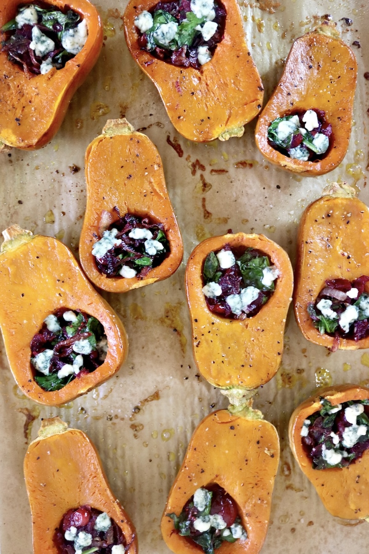 Several halved and baked honeynut squash filled with cranberries and spinach, topped with bits of roquefort cheese, on a a prchment-lined sheet pan.