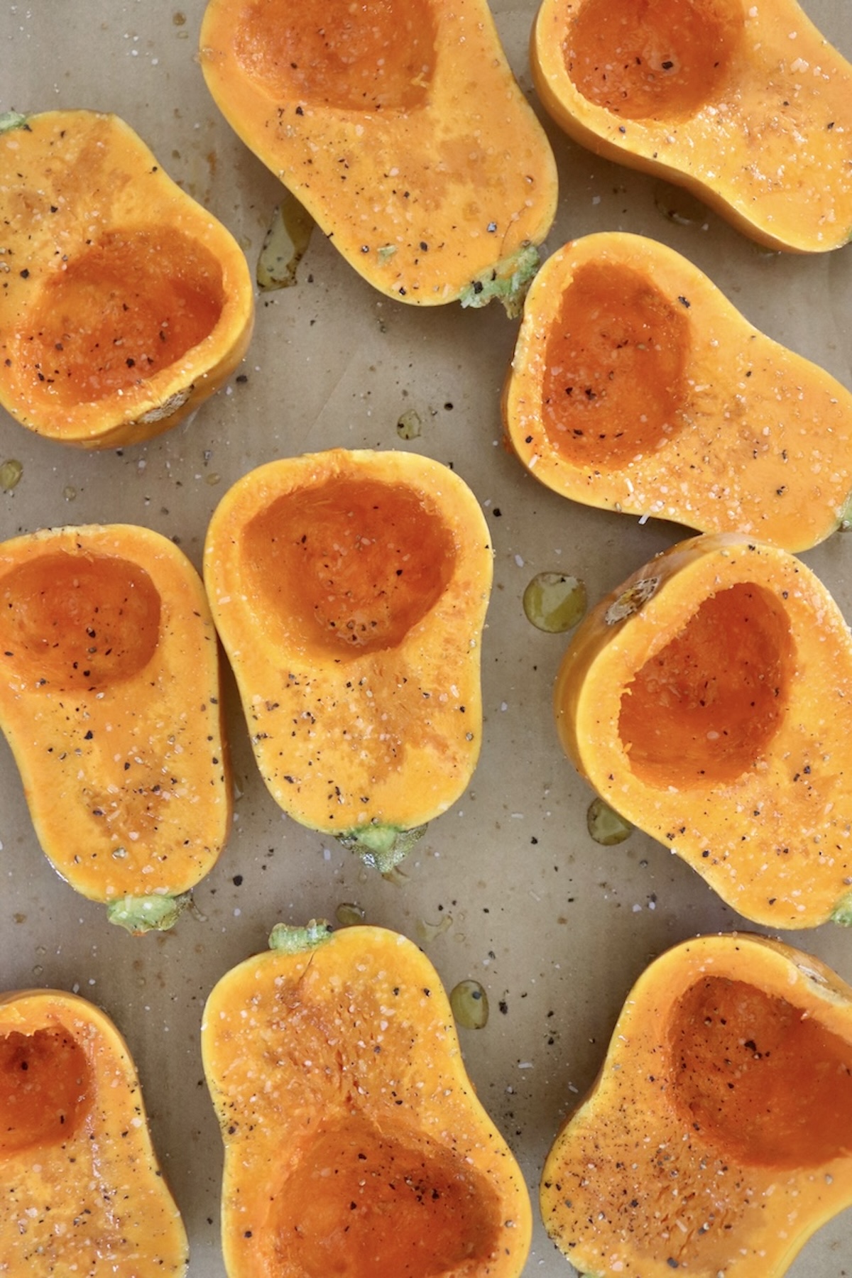 Several raw, halved honeynut squash, round side down, on a a prchment-lined sheet pan, with olive oil, salt and pepper.