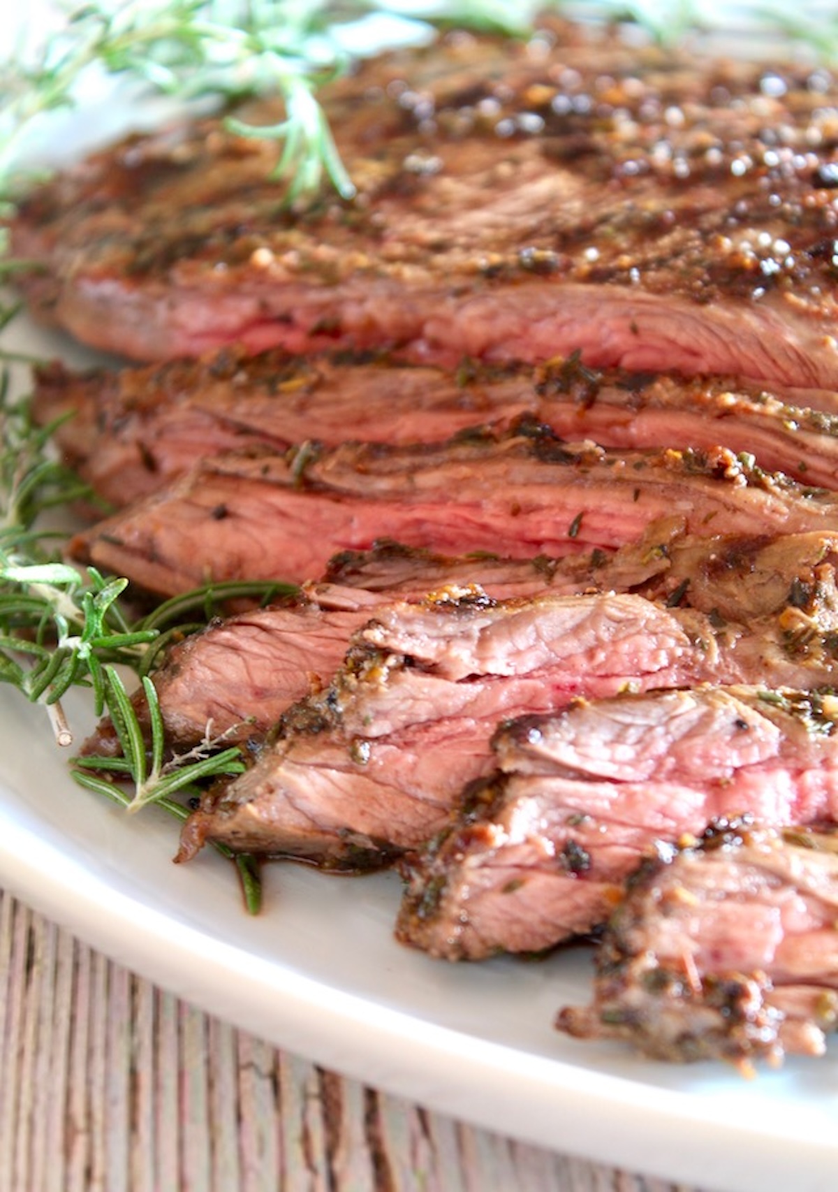 Mediterranean-Marinated-Flank-Steak Sliced marinated Flank steak on a white platter with fresh rosemary.