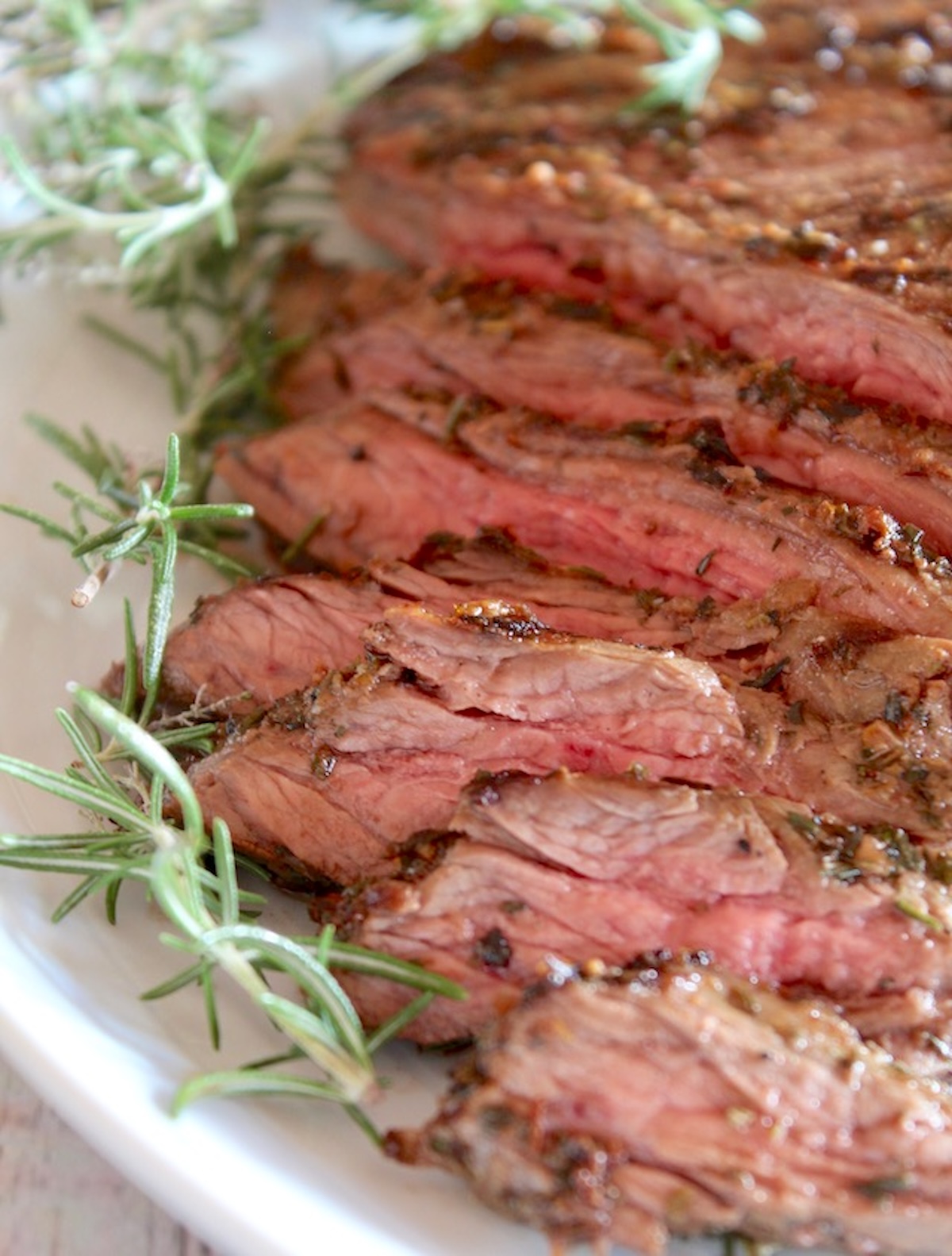 Mediterranean-Marinated-Flank-Steak Sliced marinated Flank steak on a white platter with fresh rosemary.