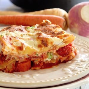 One slice of root vegetable lasagna on a cream-colored plate with a carrot and turnip behind it.