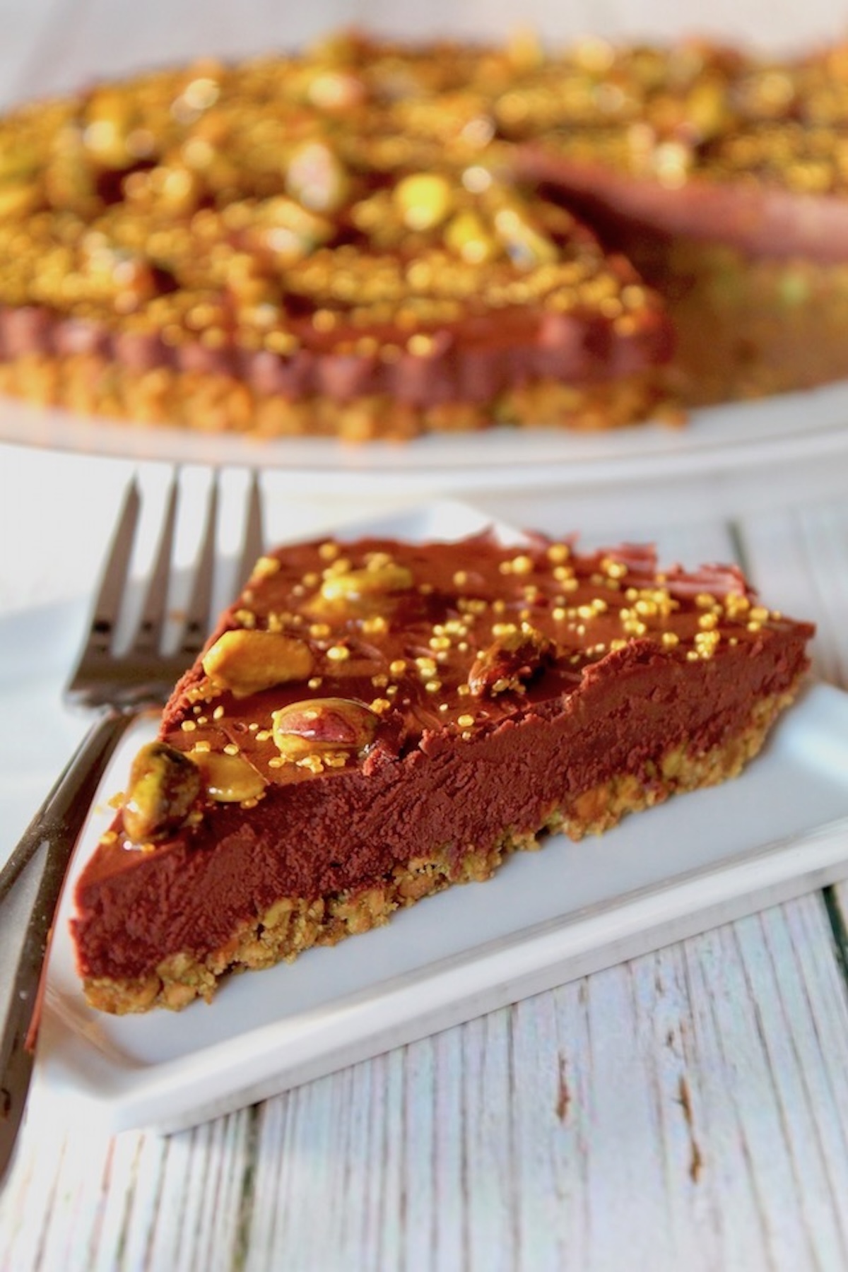 Chocolate-Tart-with-Pistachio-CrustCrust One slice of chocolate tart with pistachio crust on a white plate with whole tart behind it.