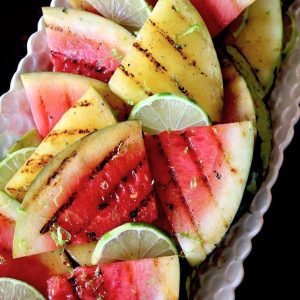 Several grill-marked triangles of yellow and red watermelon on a white narrow platter with lime wedges.