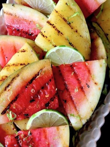 Several grill-marked triangles of yellow and red watermelon on a white narrow platter with lime wedges.