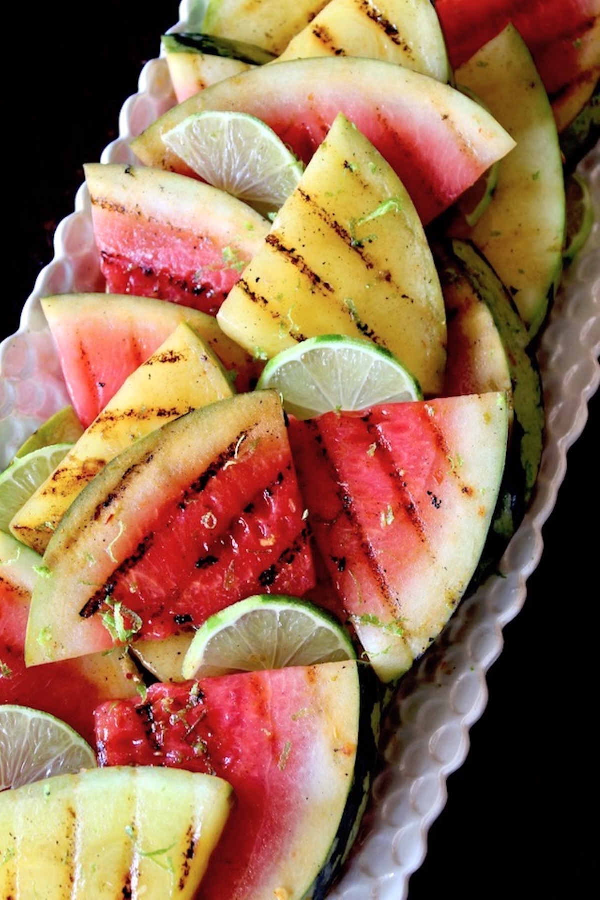 Lime-Chile-Grilled-Watermelon Several grill-marked triangles of yellow and red watermelon on a white narrow platter with lime wedges.