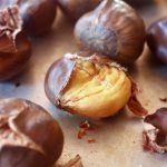 Several roasted chestnuts on a sheet pan and one with skin and shell almost all the way off.