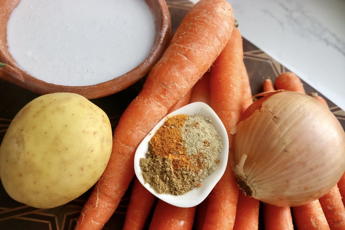 Coconut-Spiced-Carrot-Soup Ingredients for creamy carrot soup with coconut, including carrots, potato, onion, coconut milk and a small dish with cardamom, turmeric and cumin.