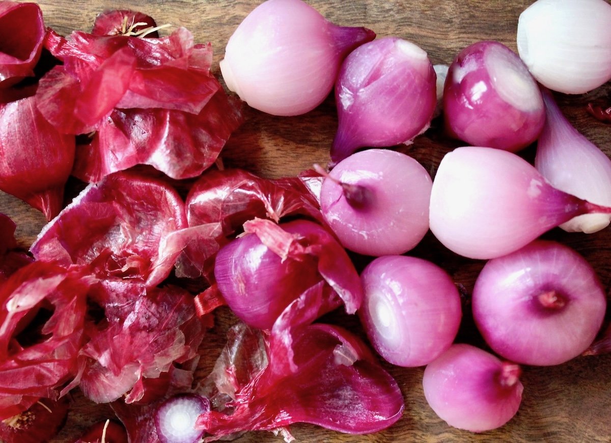 How-to-Peel-Pearl-Onions Peeled purple pearl onions with peels on dark wood cutting board.