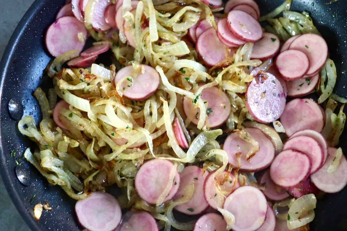 Caramelized onions and small sliced potatoes in a skillet with tongs.