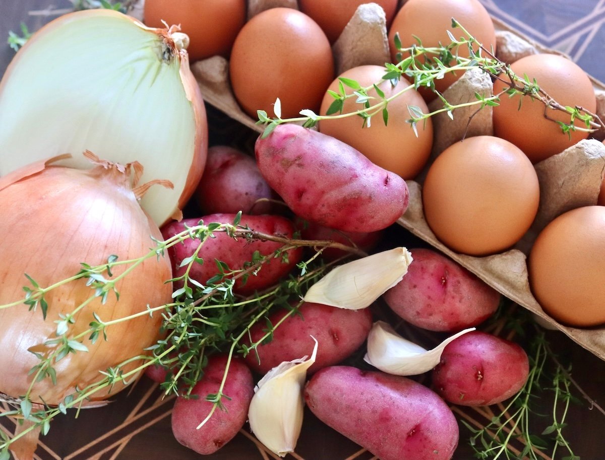 Ingredients for egg white frittata on a cutting board, including an onion, brown eggs, small red potatoes, garlic and fresh thyme.