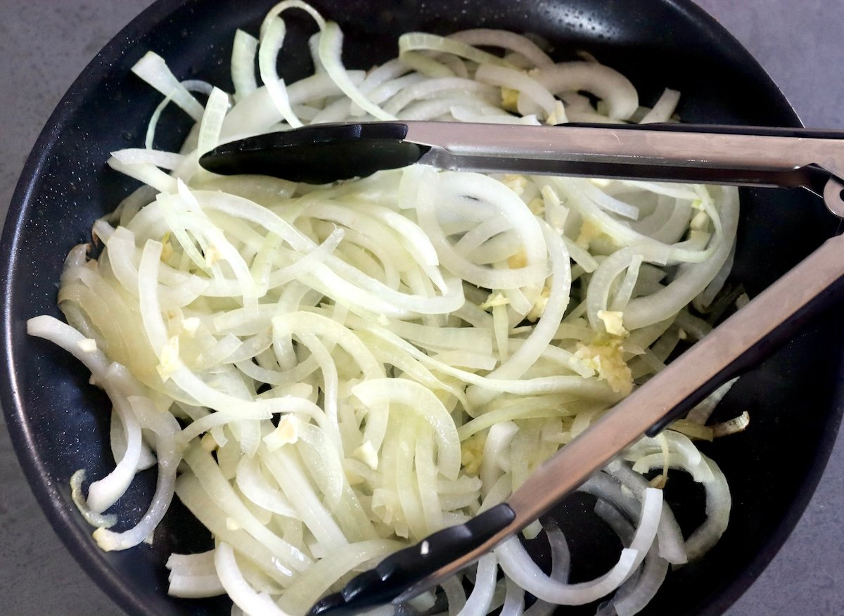 Raw sliced onions and garlic in a skillet with tongs.