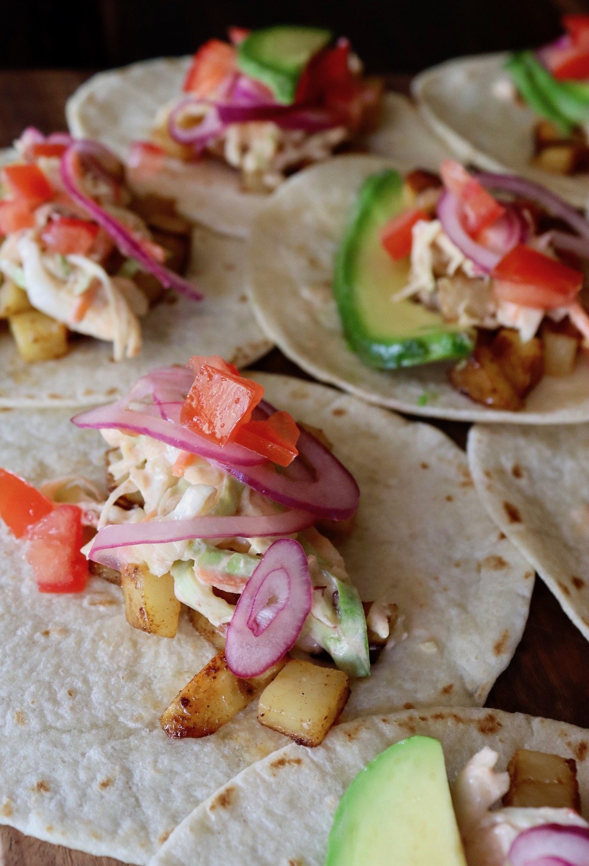 close up of a few small tortillas on a flat surface with potatoes, slaw, red onion and tomato.