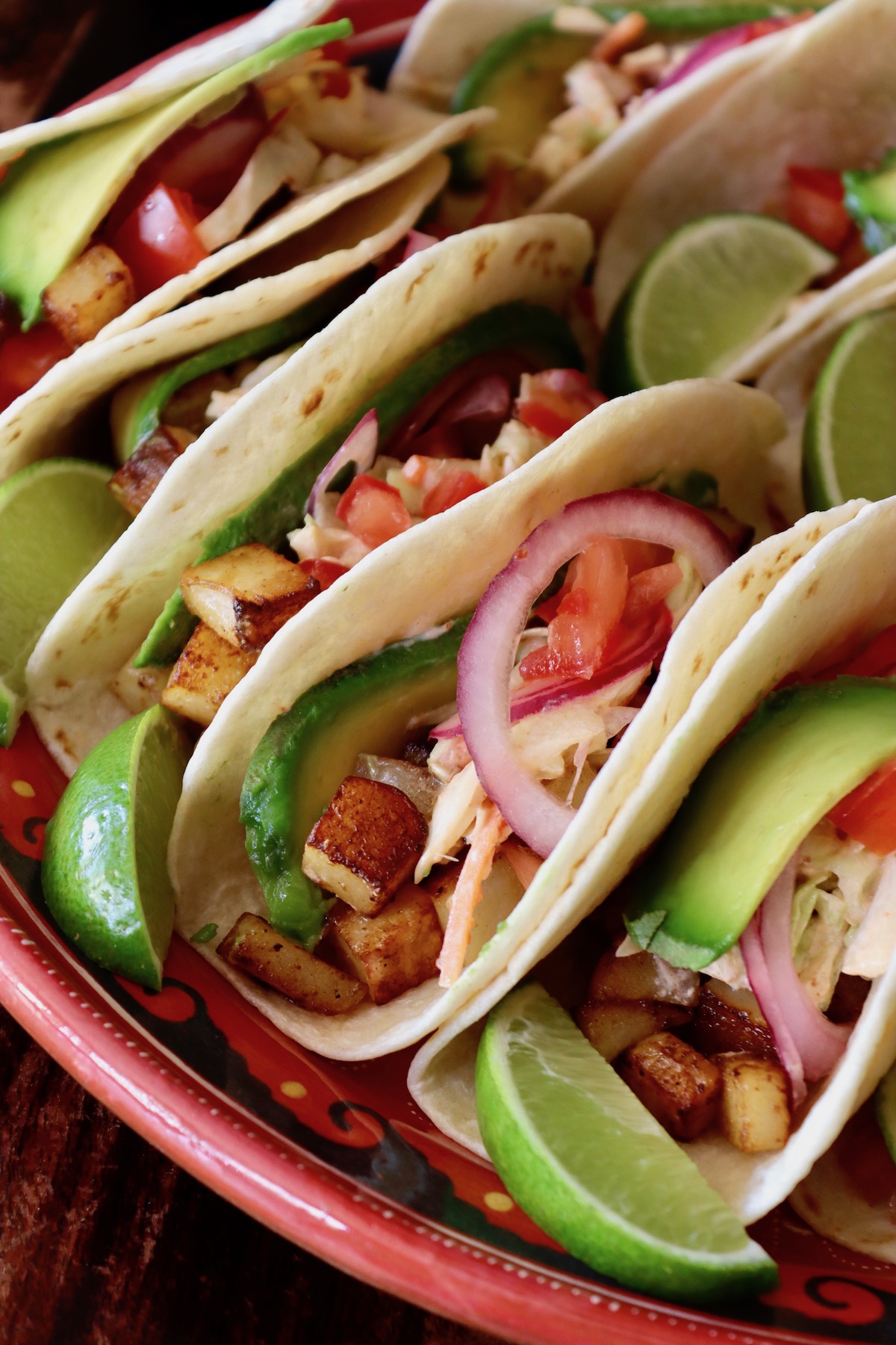 Several filled spicy potato soft tacos with avocado and lime slices on a red plate.