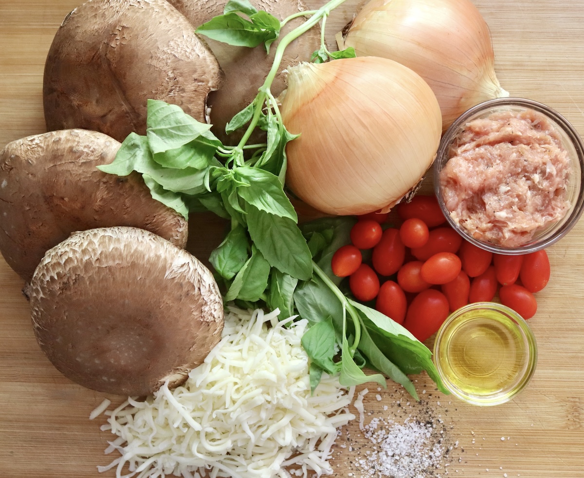 4 large portobello mushroom caps, a halved yellow onion, pile of red tear drop tomatoes, fresh basil, and pile of grated mozzarella cheese.