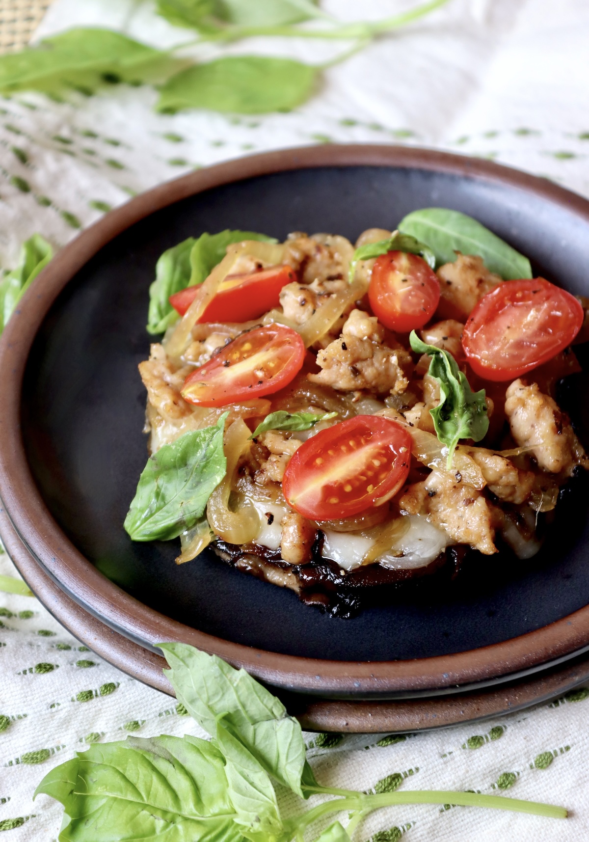 Individual portobello mushroom pizza on a black ceramic plate with caramelized onion, sausage, halved tear-drop tomatoes and fresh basil.