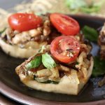 Three portions of Portobello Mushroom Pizza on a black ceramic plate, with caramelized onion, sausage, basil and tomatoes.