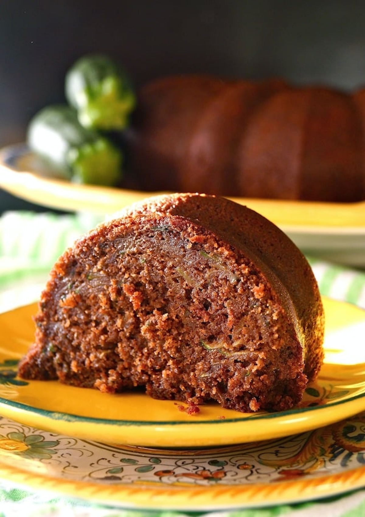 Gluten-Free-Chocolate-Zucchini-Cake-Recipe one slice of Gluten-Free Chocolate Zucchini Cake on pretty golden plate with whole cake in back ground and 2 fresh zucchini