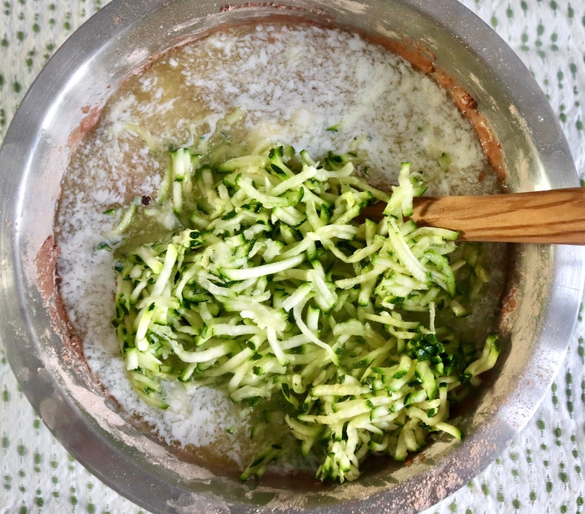 Unmixed chocolate zucchini batter with a wooden spoon in a stainless steel bowl, with pile of grated zucchini on top.