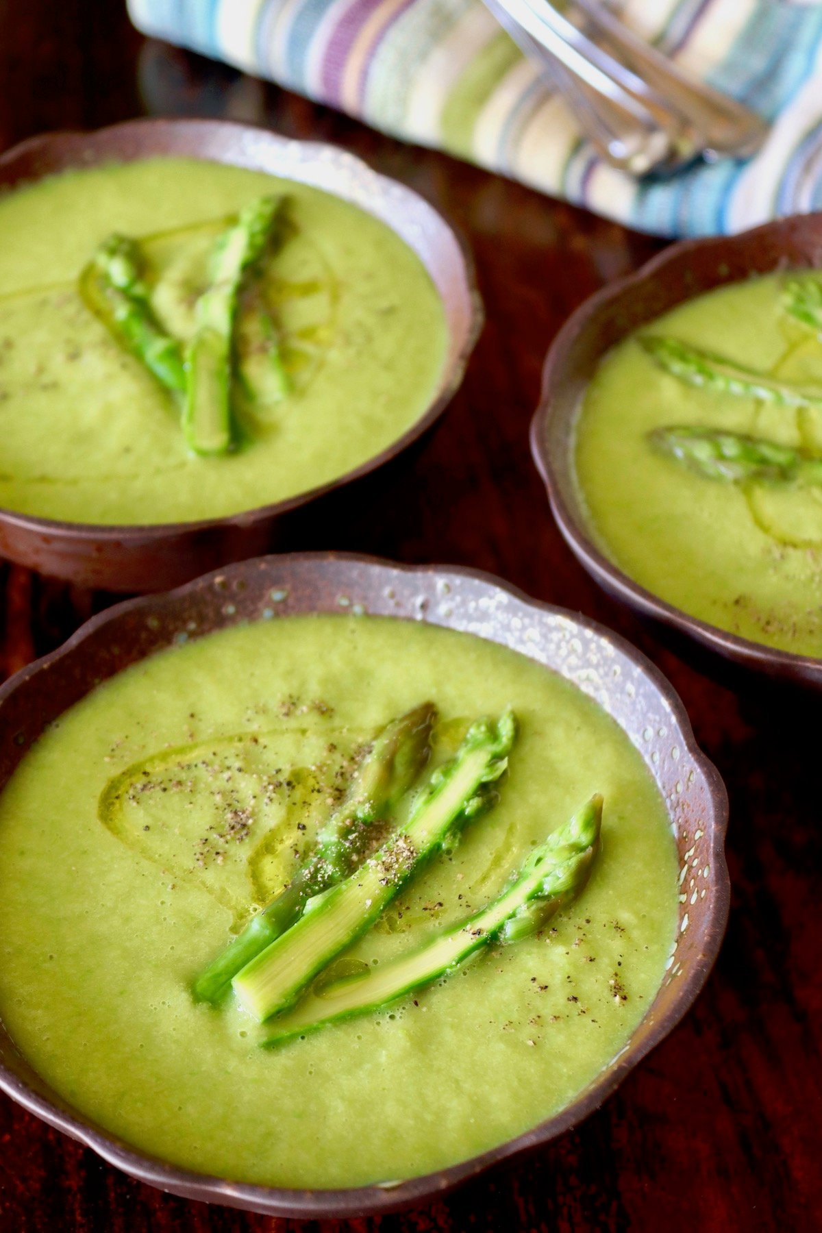 Three dark brown ceramic bowls with a fluted edge, filled with bright green asparagus soup with three asparagus spears on top.