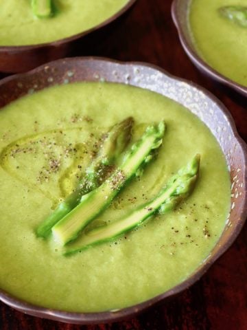 Dark brown ceramic bowl filled with bright green asparagus soup with three asparagus spears on top.