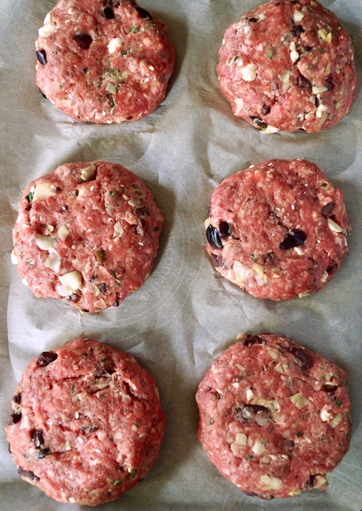 Six raw, shaped beef burgers with black olives and feta cheese.
