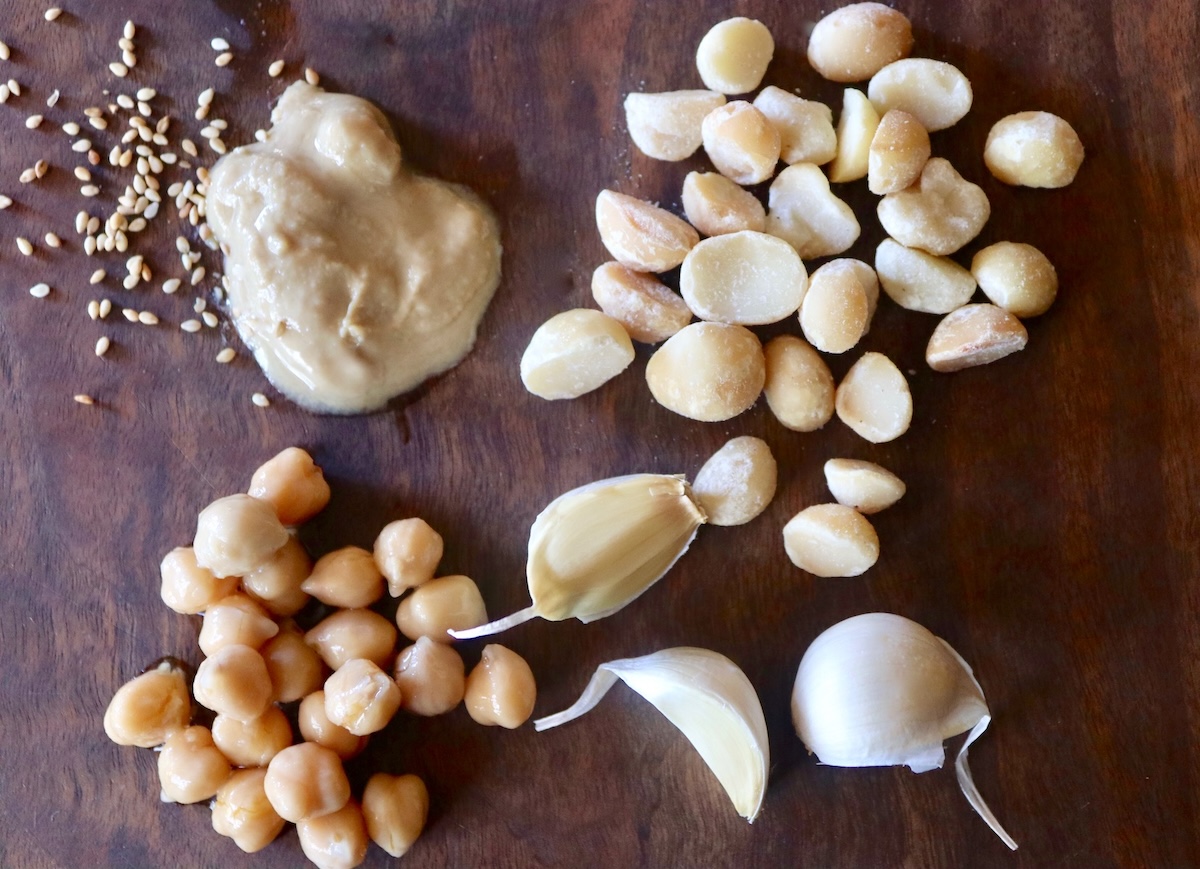 Roasted-Macadamia-Nut-Hummus-Recipe Macadamia nuts, garlic cloves, tahini and garbanzo beans on a dark wood cutting board.