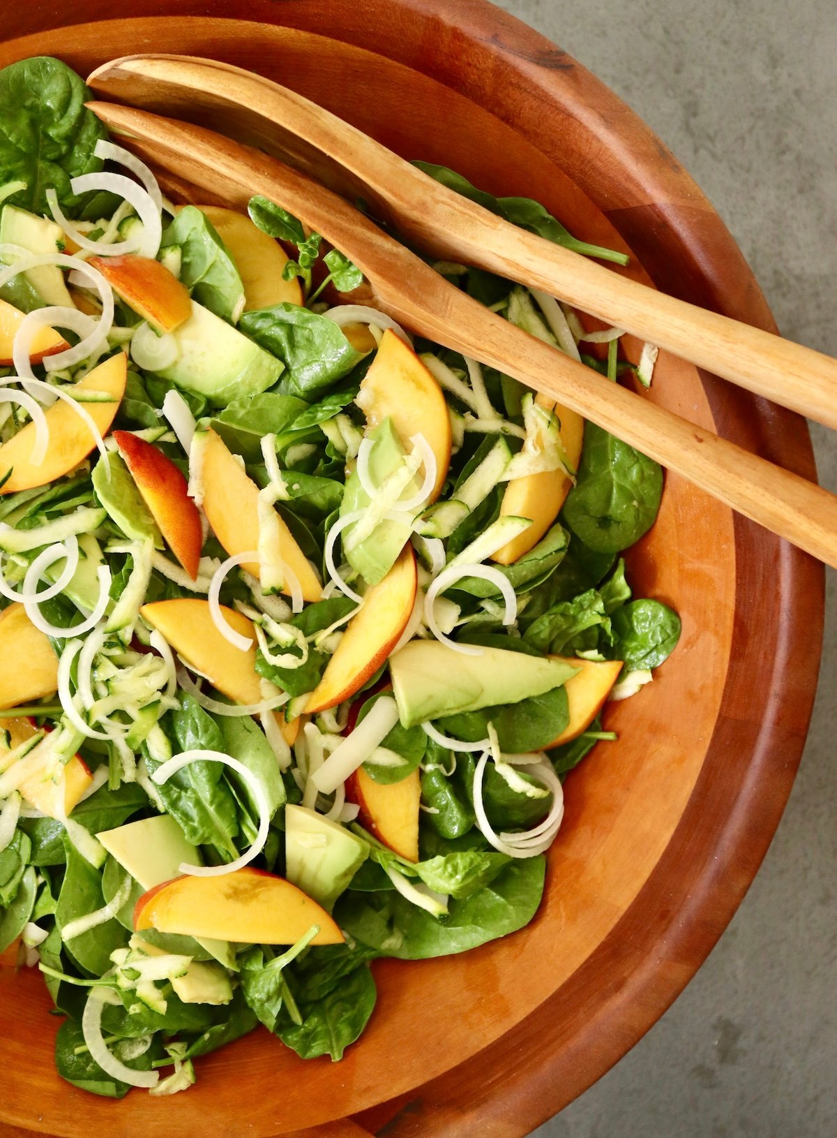 Large wooden salad bowl with avocado, peach and onion slices over baby spinach leaves, with wooden servers.