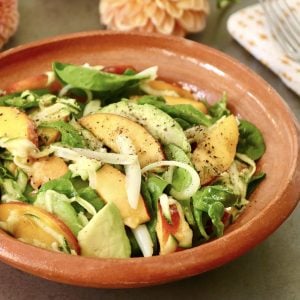 Terra cotta bowl with peach avocado salad and fresh flowers behind it.
