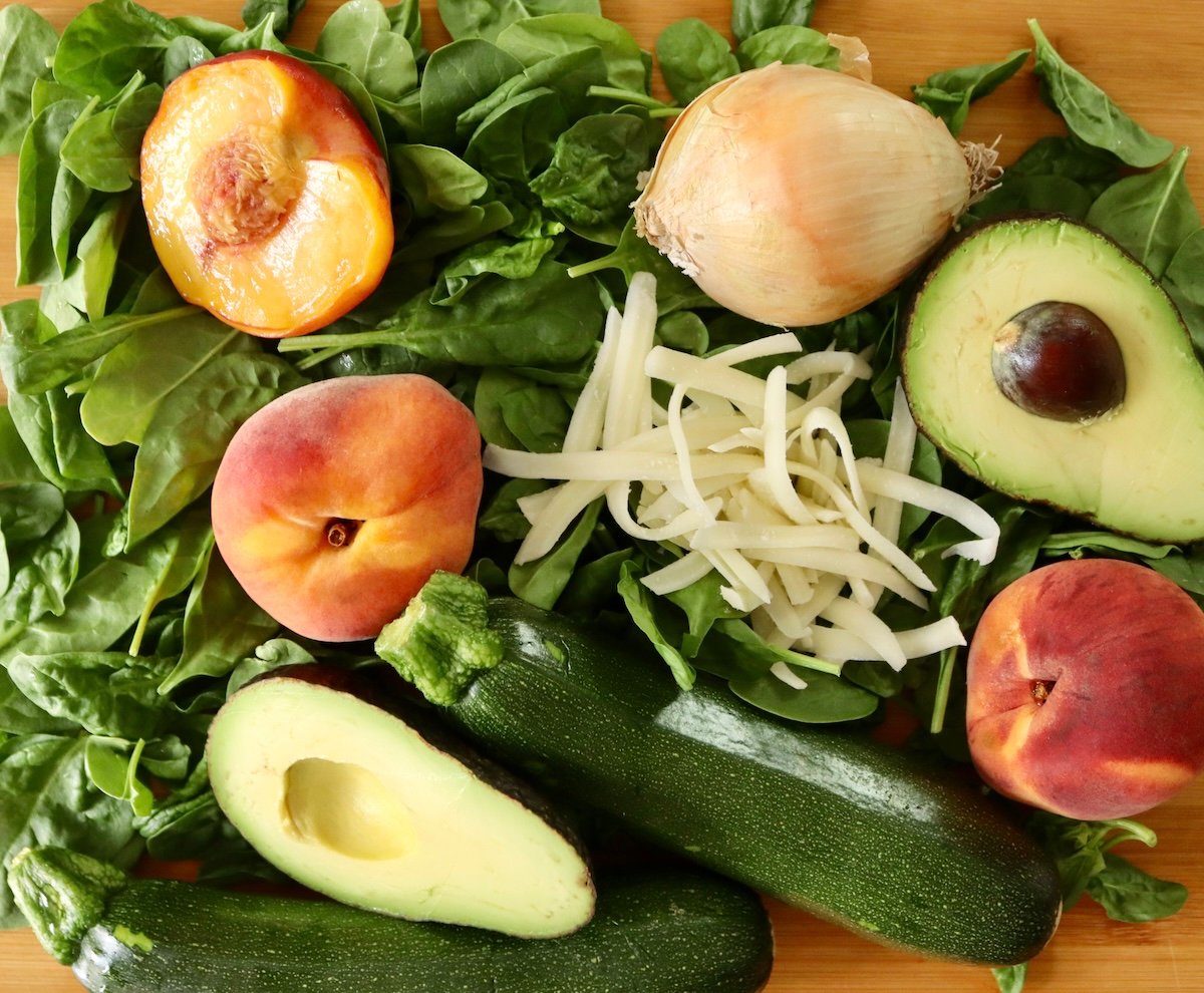 Ingredients for summer peach salad including spinach leaves, whole zucchini, avocados halves, grated mozzarella and peaches.