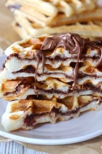 Stack of waffles cut in half with Nutella inside each one and dripping from the top, on a white plate.