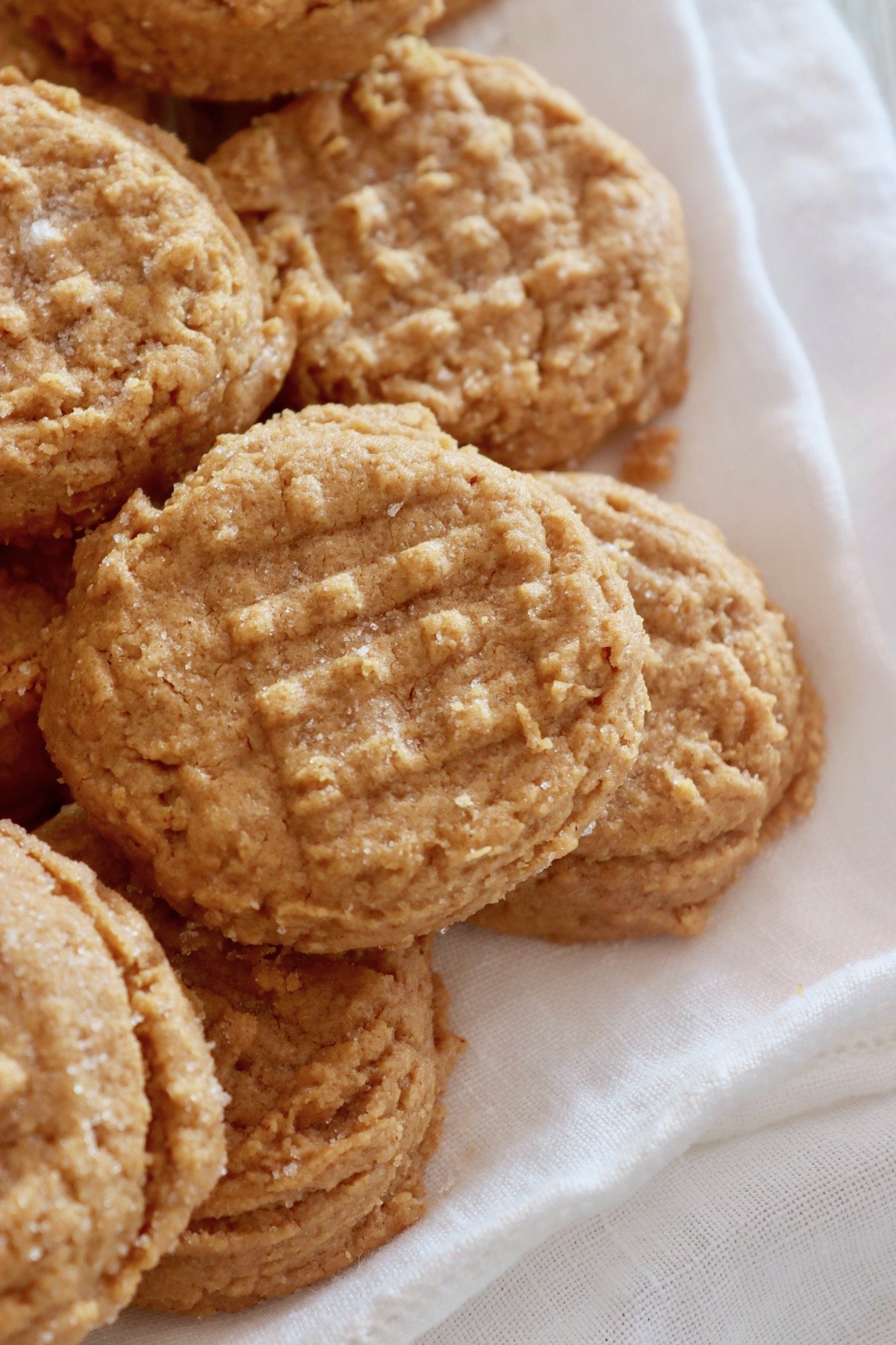 Pile of peanut butter cookies with a fork indentation in each one on white cloth.