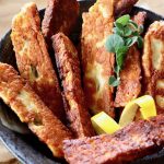 Black ceramic bowl filled with Halloumi Fries with a sprig of oregano and a lemon rind twist.