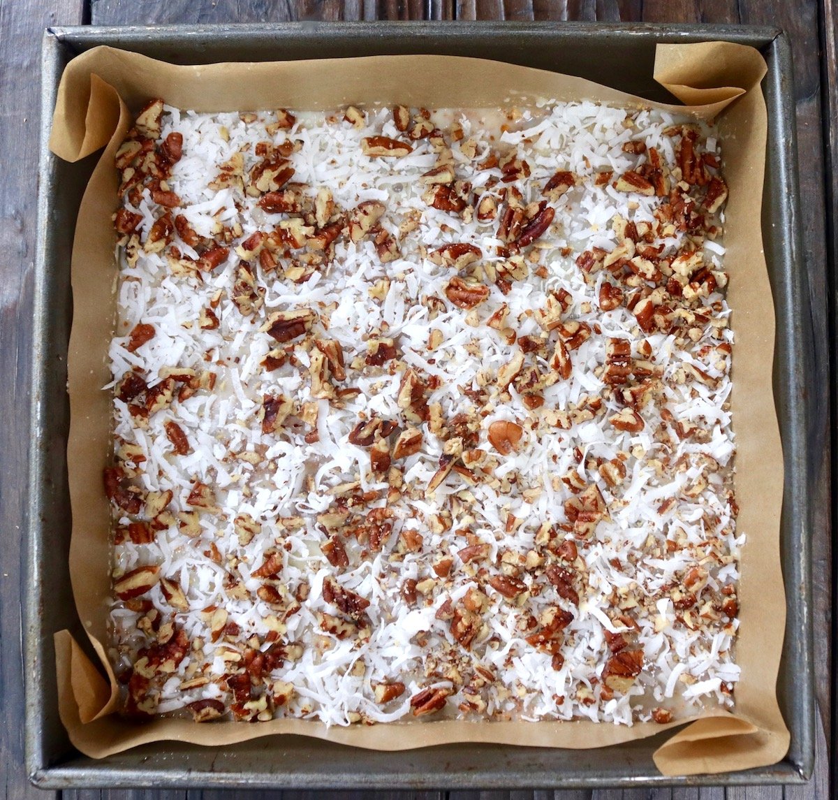 Coconut and pecan pieces in a parchment paper-lined square baking pan.