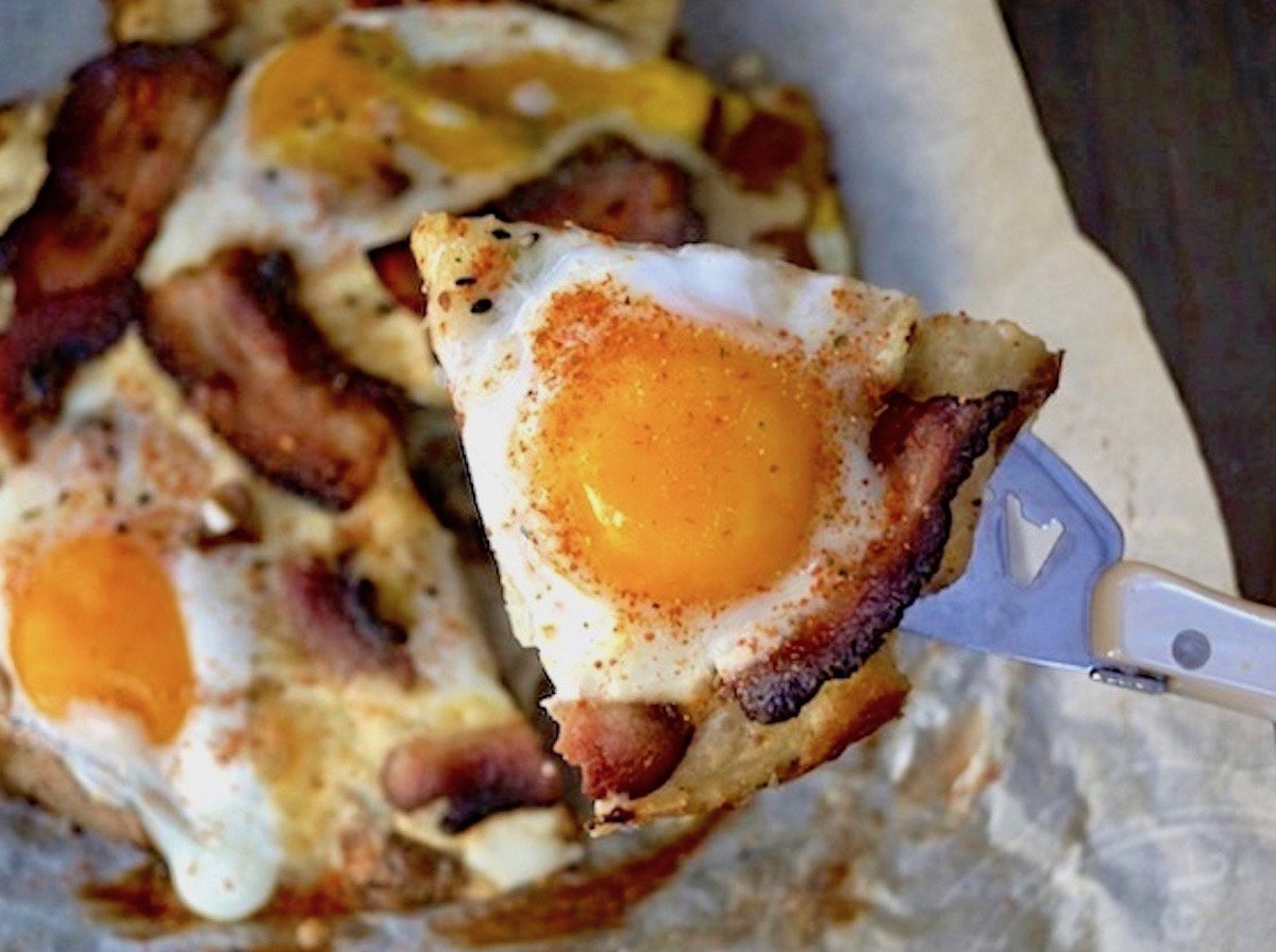 One slice of bacon and egg smashed potato pizza being held above the rest of the pizza.
