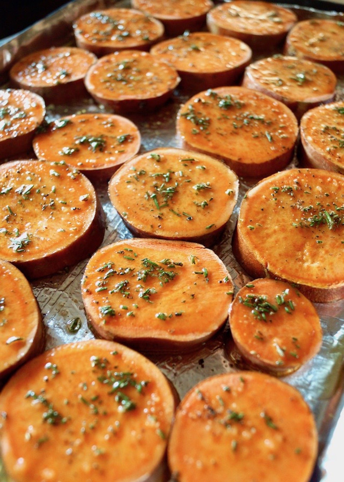 Several slices of sweet potatoes on a foil-lined baking sheet with oil and finely chopped rosemary.