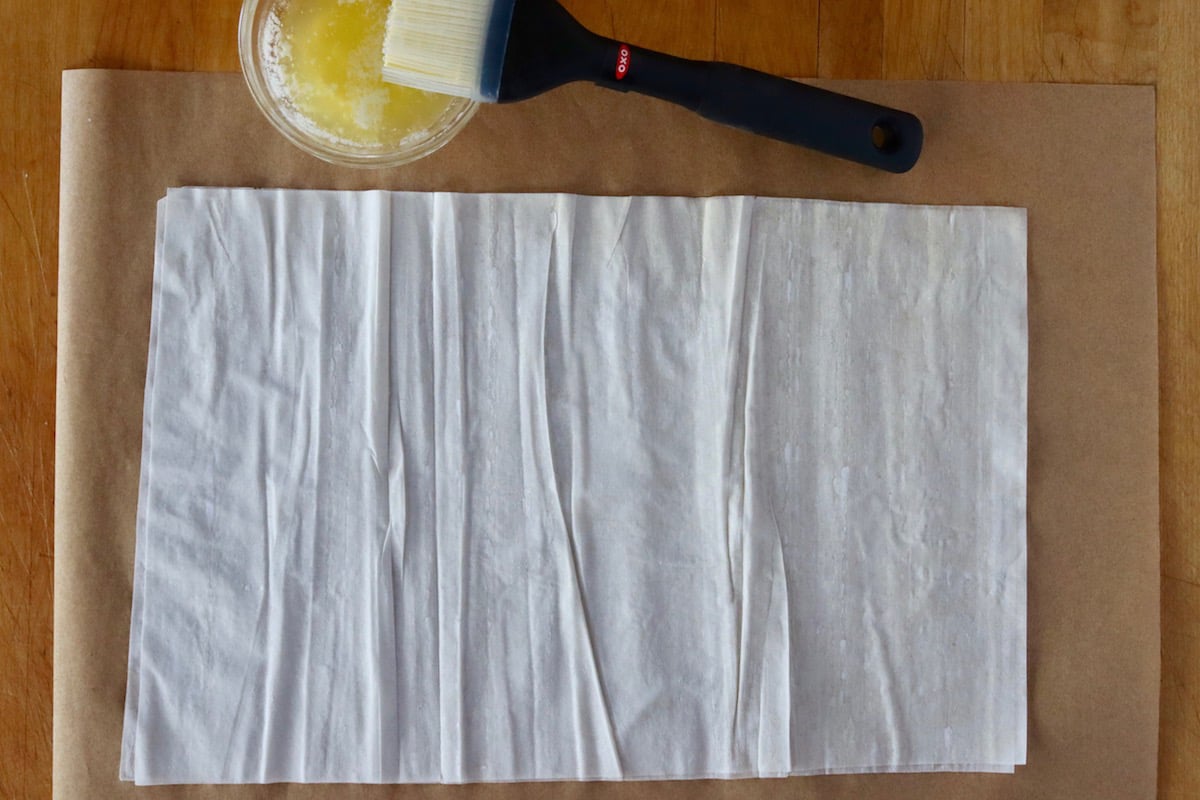 Two sheets of phyllo dough on parchment paper with a tiny bowl of melted butter and a pastry brush.