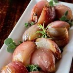 Roasted red onions, quartered, on a white narrow plate with fresh herbs.